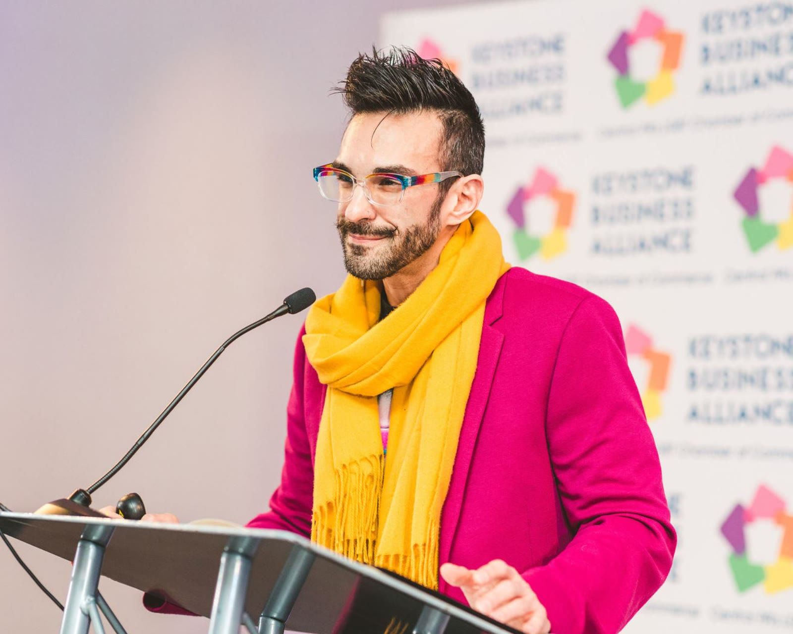 Peter Andrew Danzig speaking at a lectern in a magenta coat and yellow scarf, wearing rainbow patterned glasses, in front of a Keystone Business Alliance backdrop.