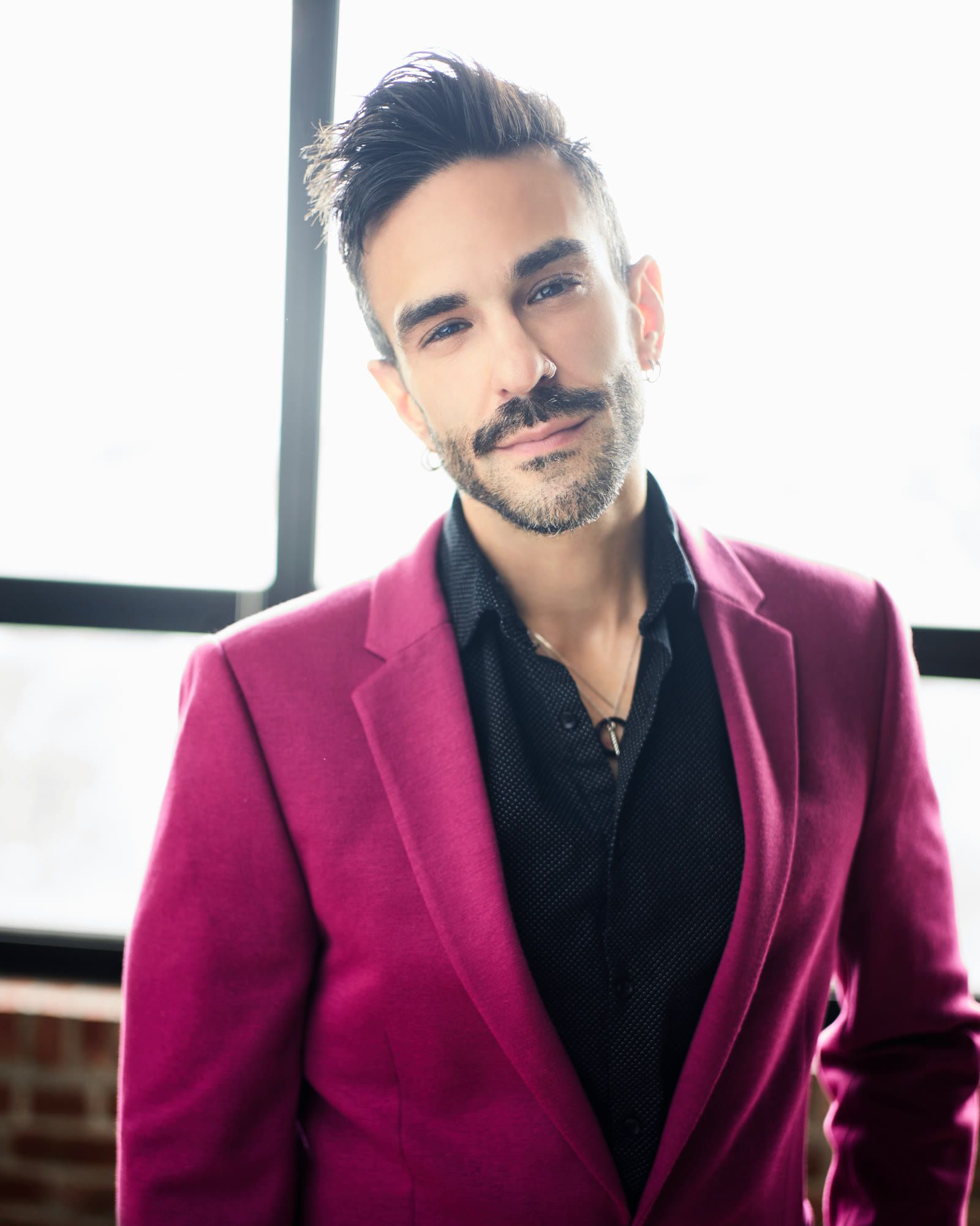 Peter Andrew Danzig standing in a fuchsia blazer over a dark open collar shirt, looking directly at the camera with a warm smile, in front of a bright window.