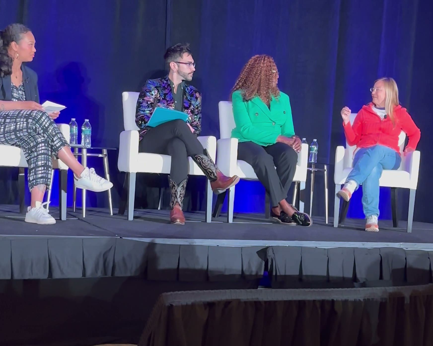 Peter Andrew Danzig seated second from left on a conference main stage with three other panelists, wearing an iridescent floral blazer and holding a teal notebook, against a deep blue backdrop.