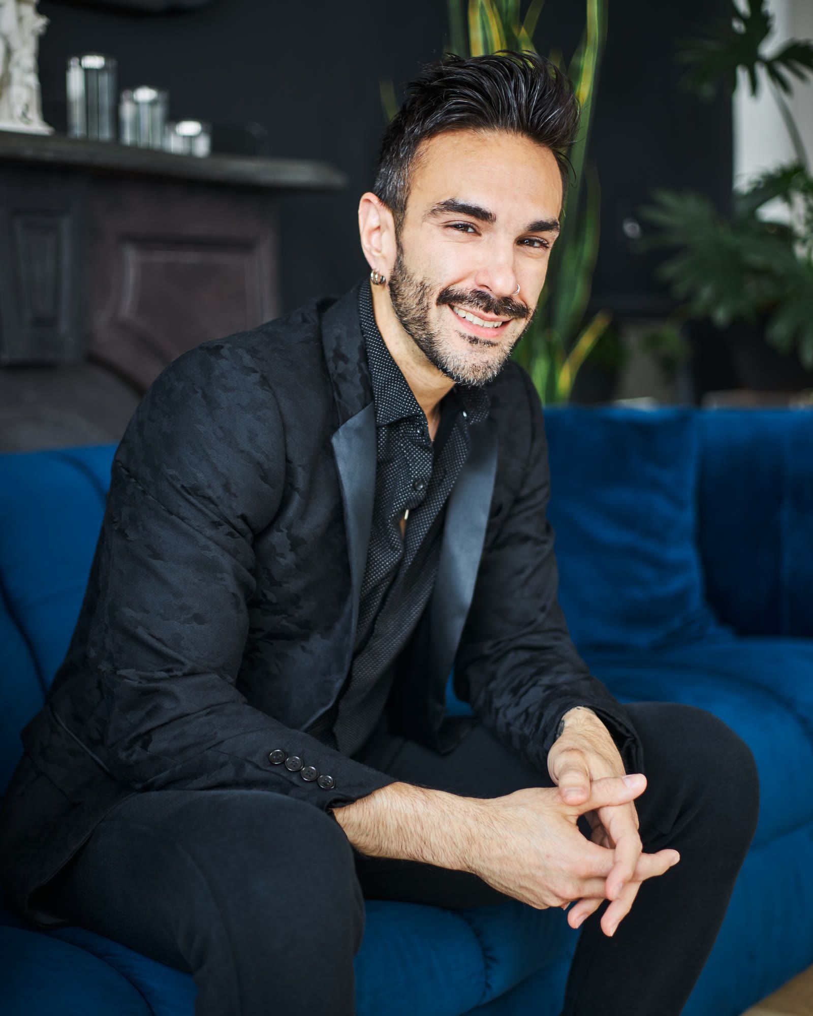 Peter Andrew Danzig seated on a blue velvet sofa in a black jacket, hands loosely clasped, with a warm open smile.