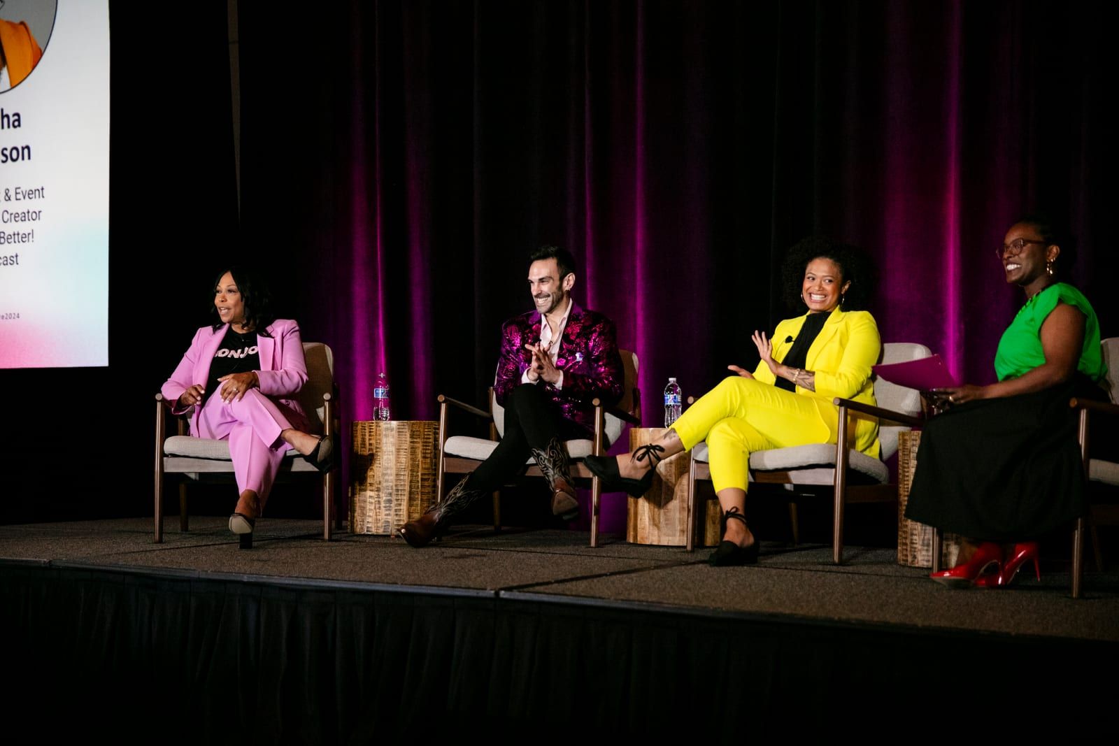 Peter Andrew Danzig seated second from left on a conference panel of four, in a magenta velvet blazer and boots, laughing and gesturing mid-answer, against a magenta curtain.