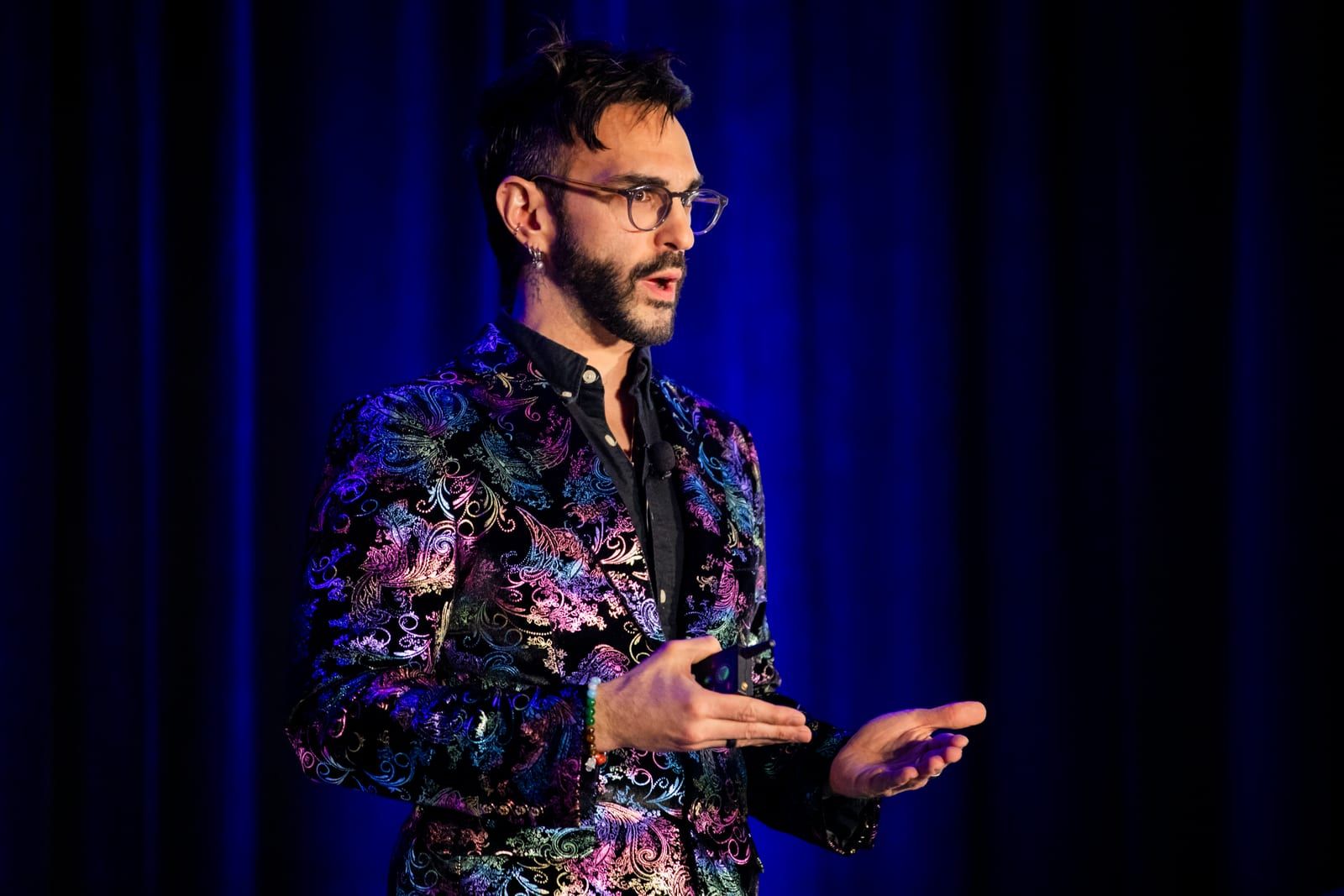 Peter Andrew Danzig speaking on a conference stage in an iridescent paisley velvet blazer and glasses, both hands open in mid-gesture, a lapel microphone clipped to their collar, against a deep blue curtain.